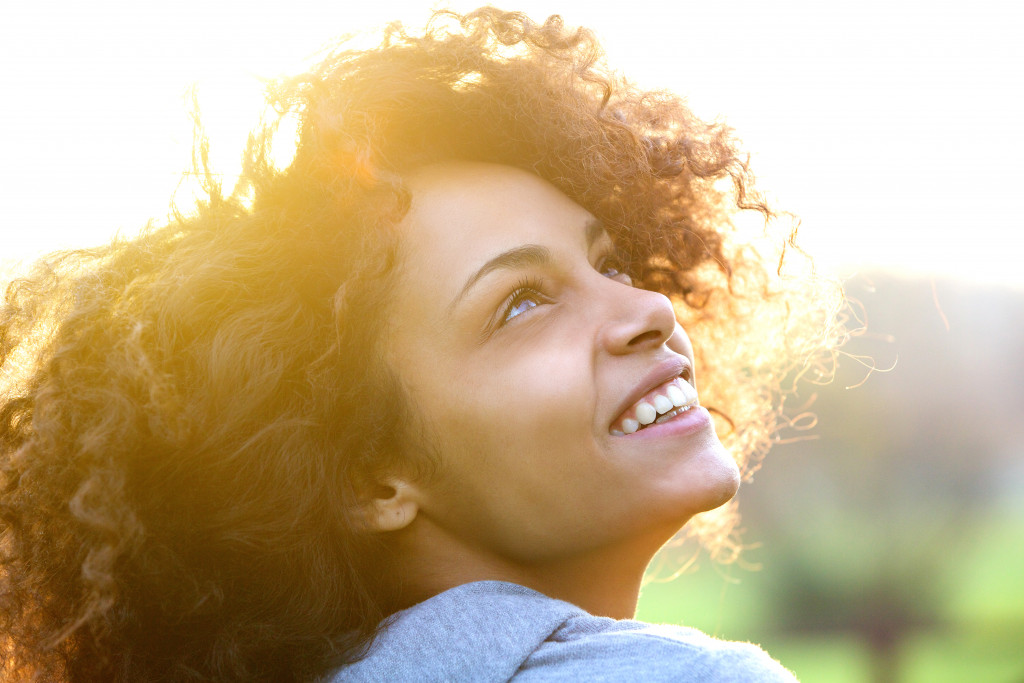 curly haired woman smiling while looking up at the sun outdoors