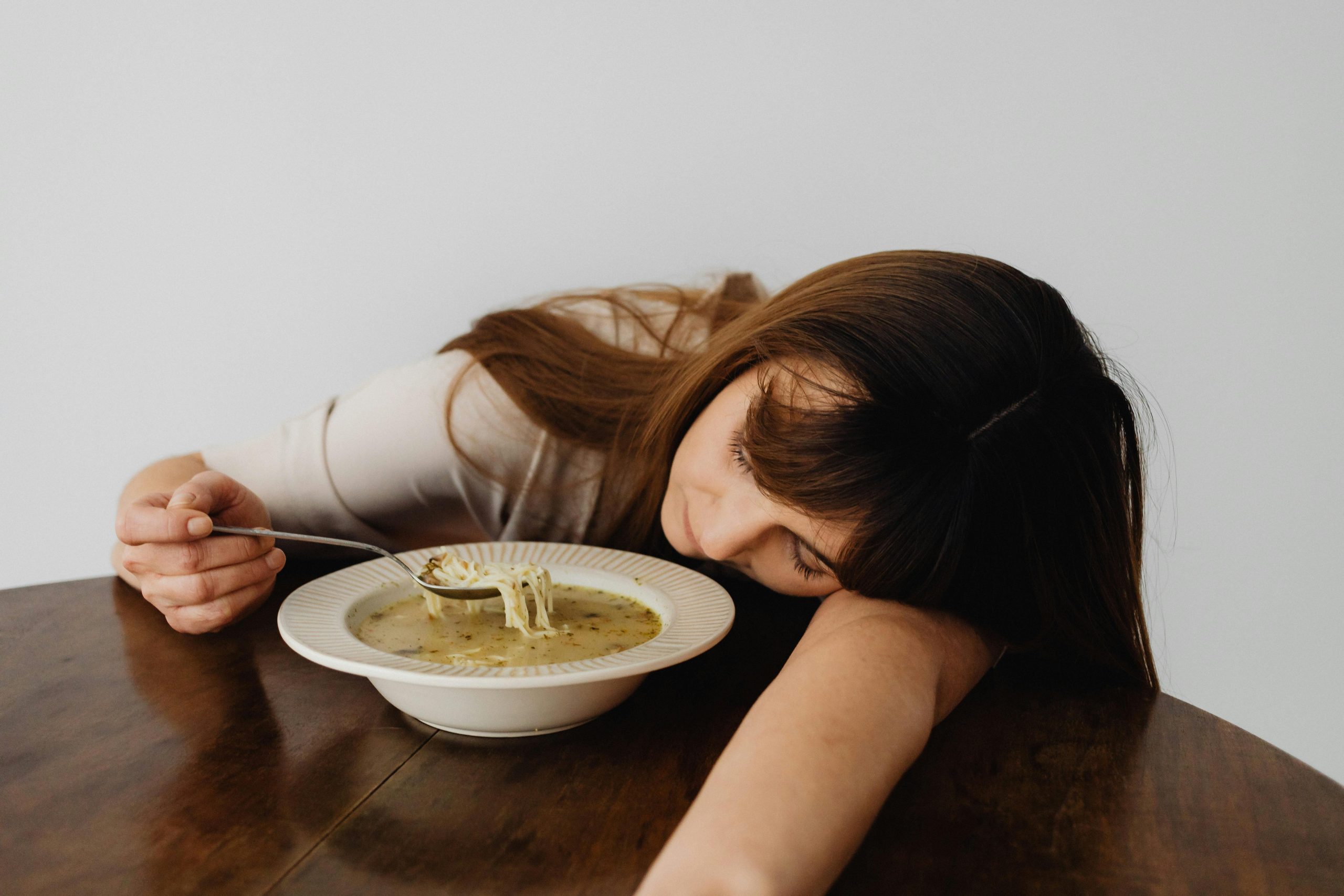 tired woman resting her head on her arm on a table, holding a spoon over a bowl of soup