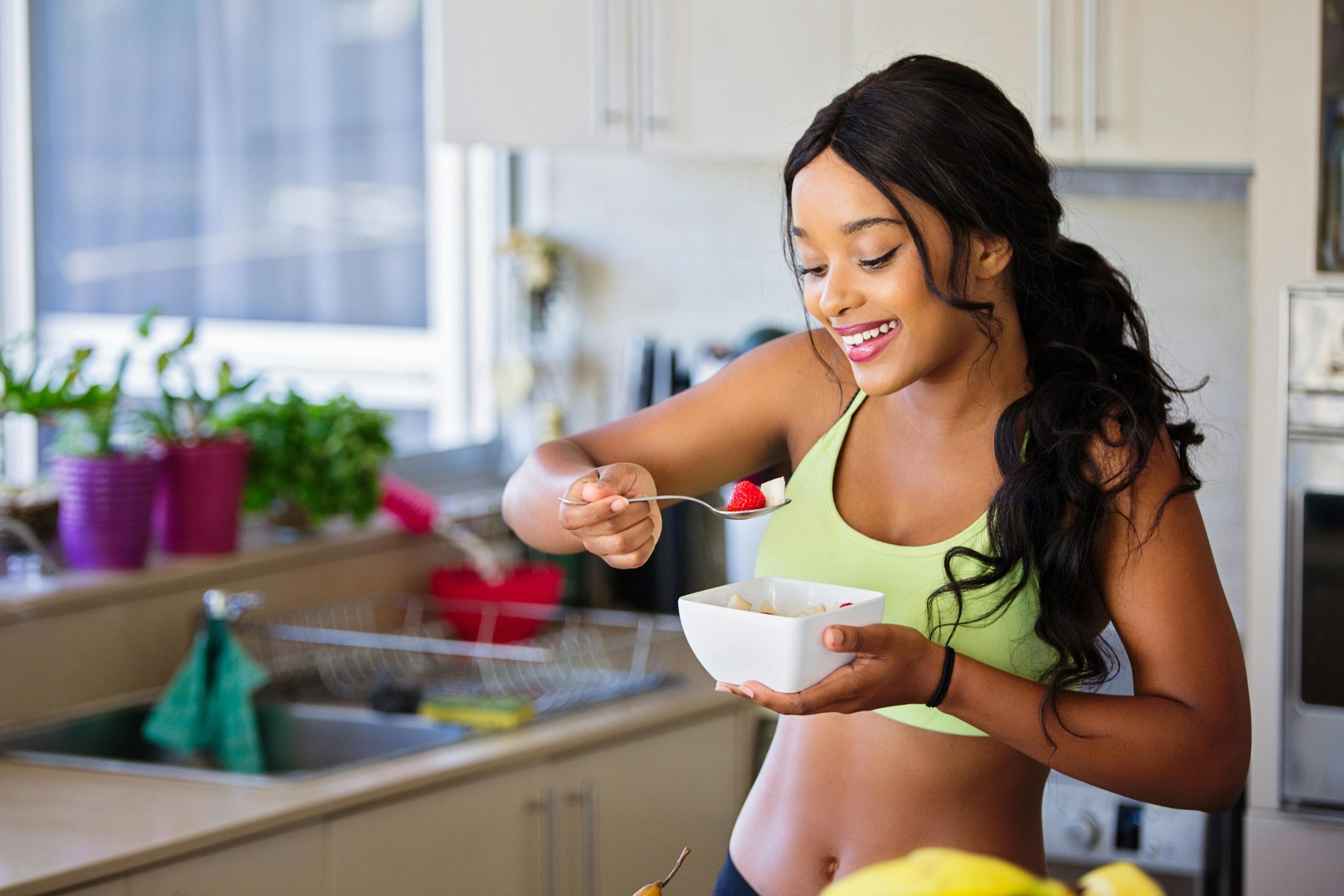 woman in workout clothes eating a bowl of fruits in a kitchen