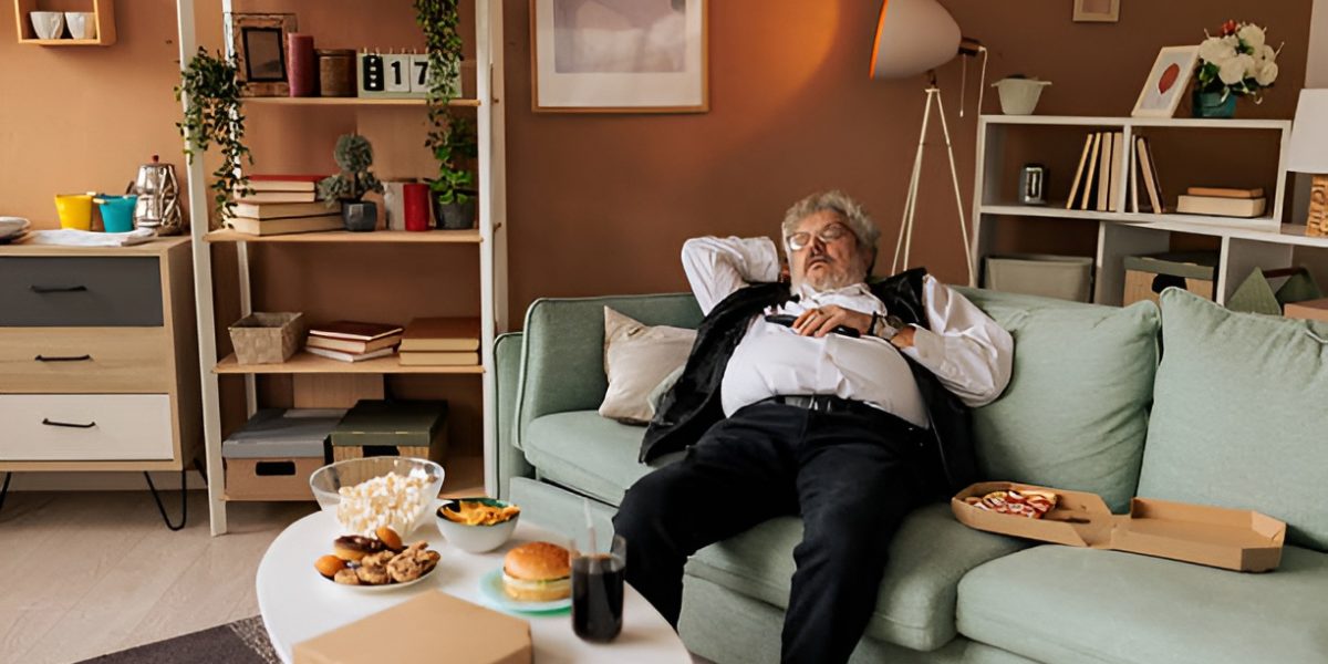 man sleeping on a couch, next to a box of pizza and a coffee table filled with comfort food