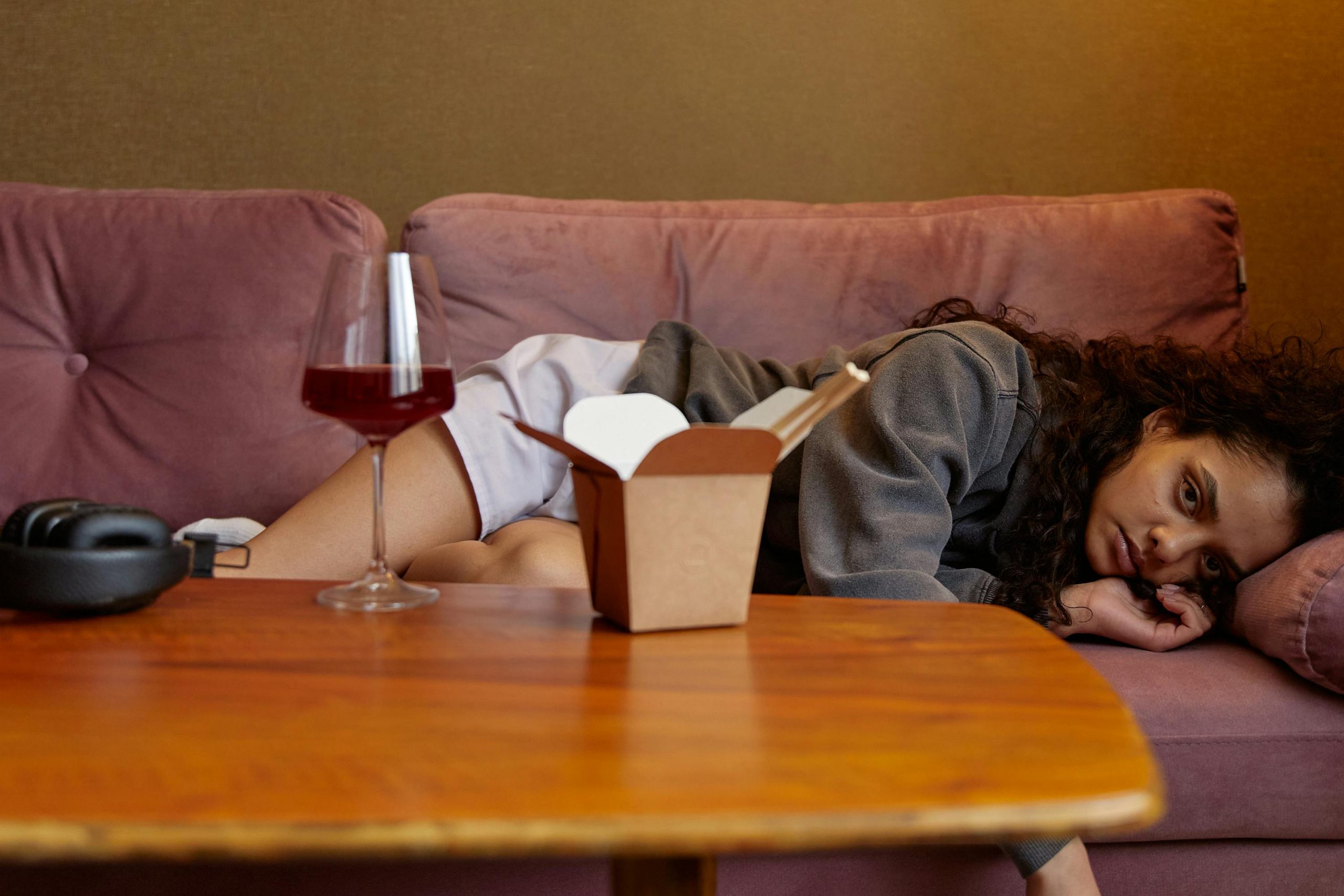 woman lying on a pink couch, next to a coffee table with a glass of wine and an open Chinese takeout box